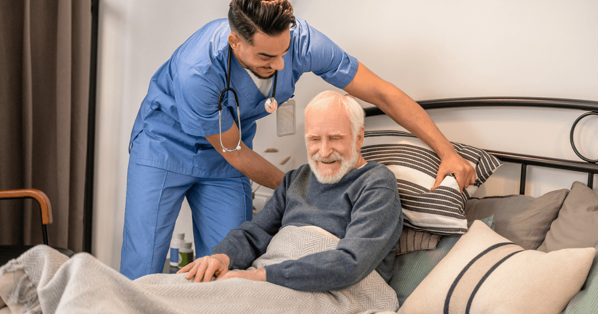 Male caregiver in scrubs adjusting older mans pillow behind him while he sits in bed.